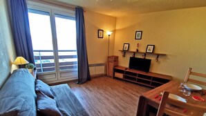 A living room with a sofa, a TV, a dining table with chairs, and a balcony with curtains. - 2-Room Cabin at the Foot of the Slopes - 4 Bunk Beds, Pets Allowed (Les Adrets-Prapoutel)