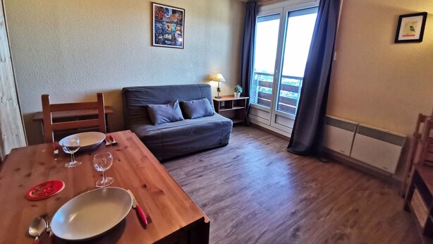 A cozy room with a wooden dining table set for two, a grey sofa, and a large window with curtains. - 2-Room Cabin at the Foot of the Slopes - 4 Bunk Beds, Pets Allowed (Les Adrets-Prapoutel)