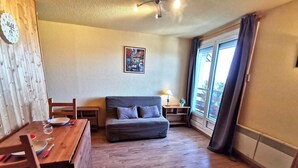 A cozy room with a wooden floor, a grey sofa, a wooden table with chairs, a small desk, and a window with curtains. - 2-Room Cabin at the Foot of the Slopes - 4 Bunk Beds, Pets Allowed (Les Adrets-Prapoutel)