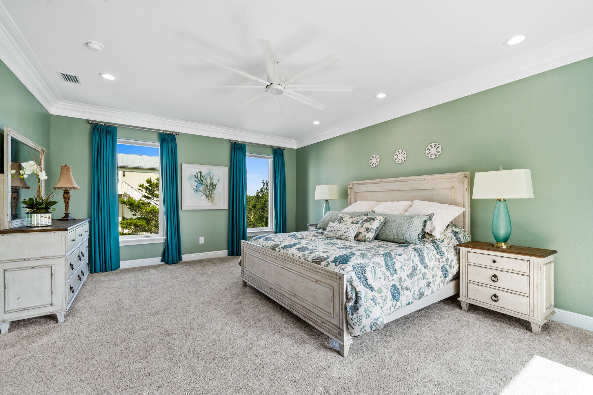 Une chambre à coucher comprenant un lit, des tables de chevet, une commode, un ventilateur de plafond et une fenêtre avec des rideaux.