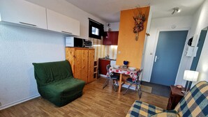 Un espacio habitable compacto con un sofá verde, un sofá a cuadros, una mesa de comedor con sillas, una kitchenette y una puerta.