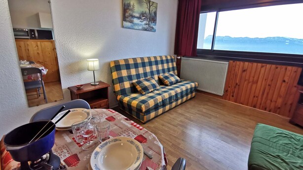Una sala de estar acogedora con un sofá de cuadros, una mesa de comedor con platos y vasos, una lámpara y vistas al exterior a través de una ventana.