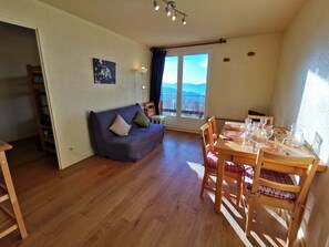 A cozy living room with a sofa, wooden dining table, chairs, and a balcony with a view. - 2 Rooms - Near Slopes - Pets Allowed (Les Adrets-Prapoutel)