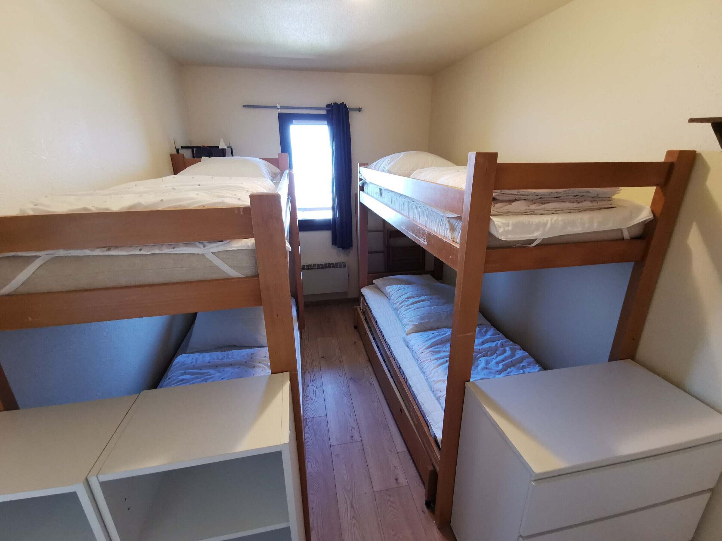A room with bunk beds, a small window with curtains, and a wooden floor.