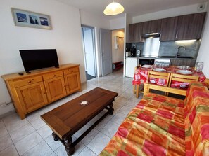 Sala de estar com um rack de madeira para a TV, uma mesa de centro e um sofá. Há uma área de cozinha com uma mesa de jantar para quatro pessoas.