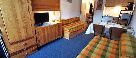 A compact room with a bed, a sofa, a television, a small table, and a kitchenette.