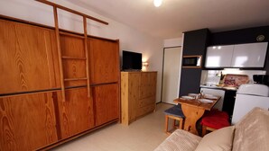 A compact living space with a wooden wardrobe, a small dining table with chairs, a sofa, and a kitchenette with a microwave and refrigerator.