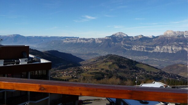 A mountain view from a balcony with a wooden railing, overlooking a valley with buildings and mountains in the distance.