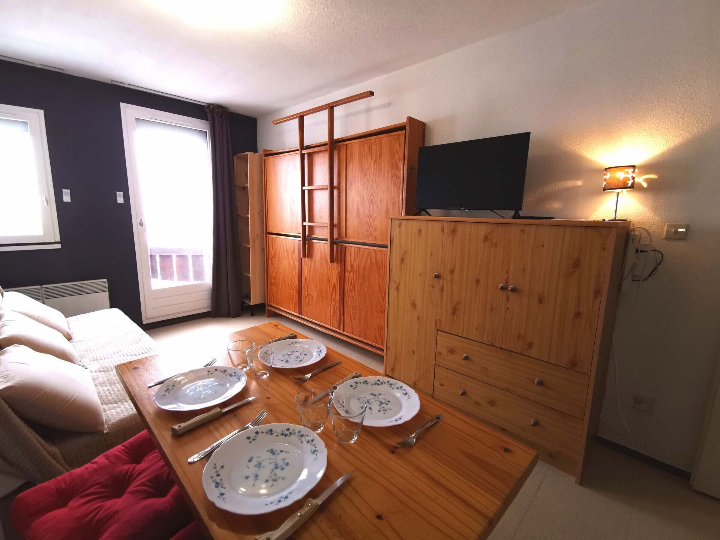 A compact room with a dining table set for four, a sofa, a TV on a stand, and a wardrobe.