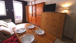 A compact room with a dining table set for four, a sofa, a TV on a stand, and a wardrobe.