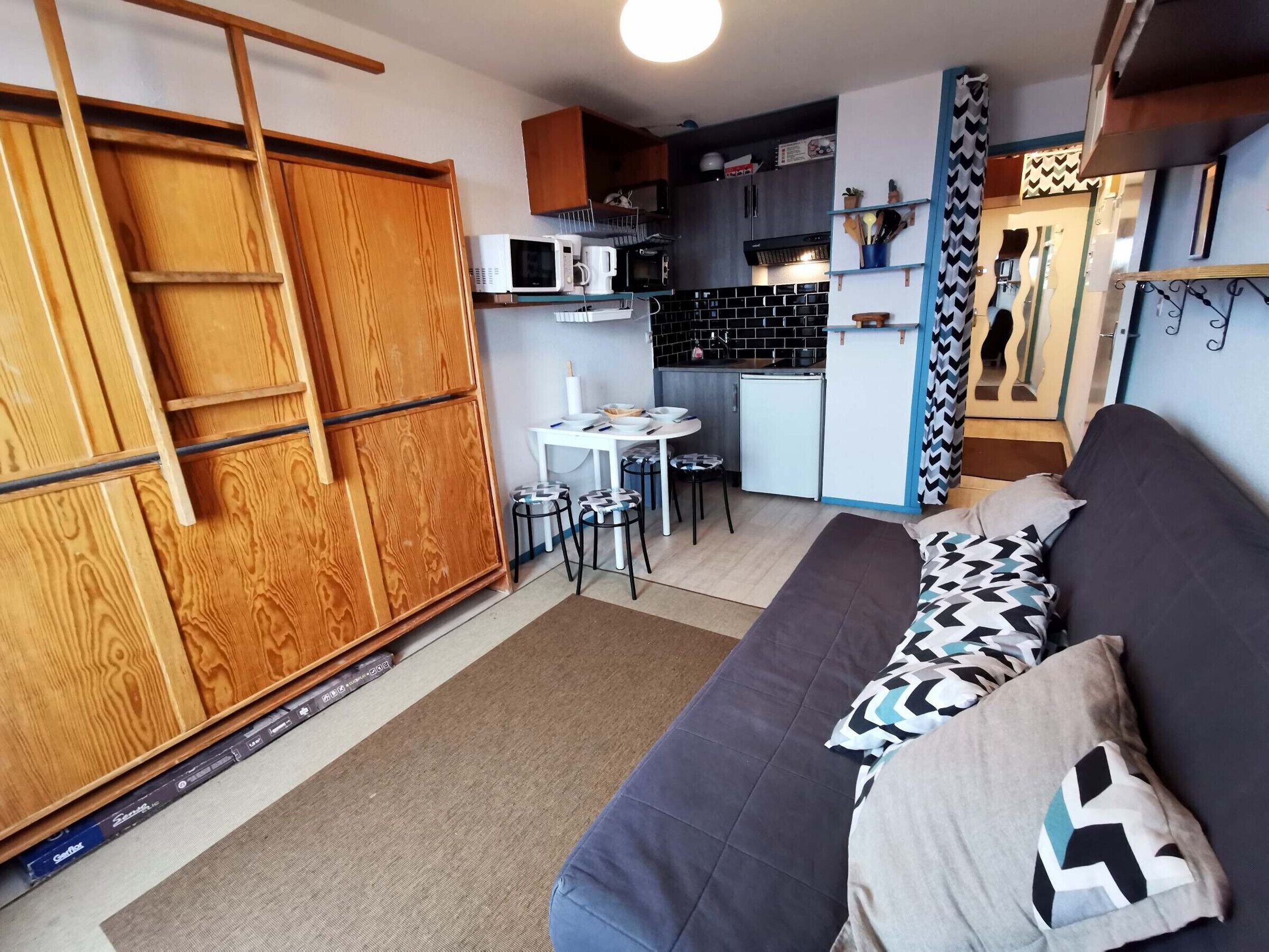 A compact living space with a kitchenette, a sofa with patterned pillows, a small dining table, and a wooden wardrobe.
