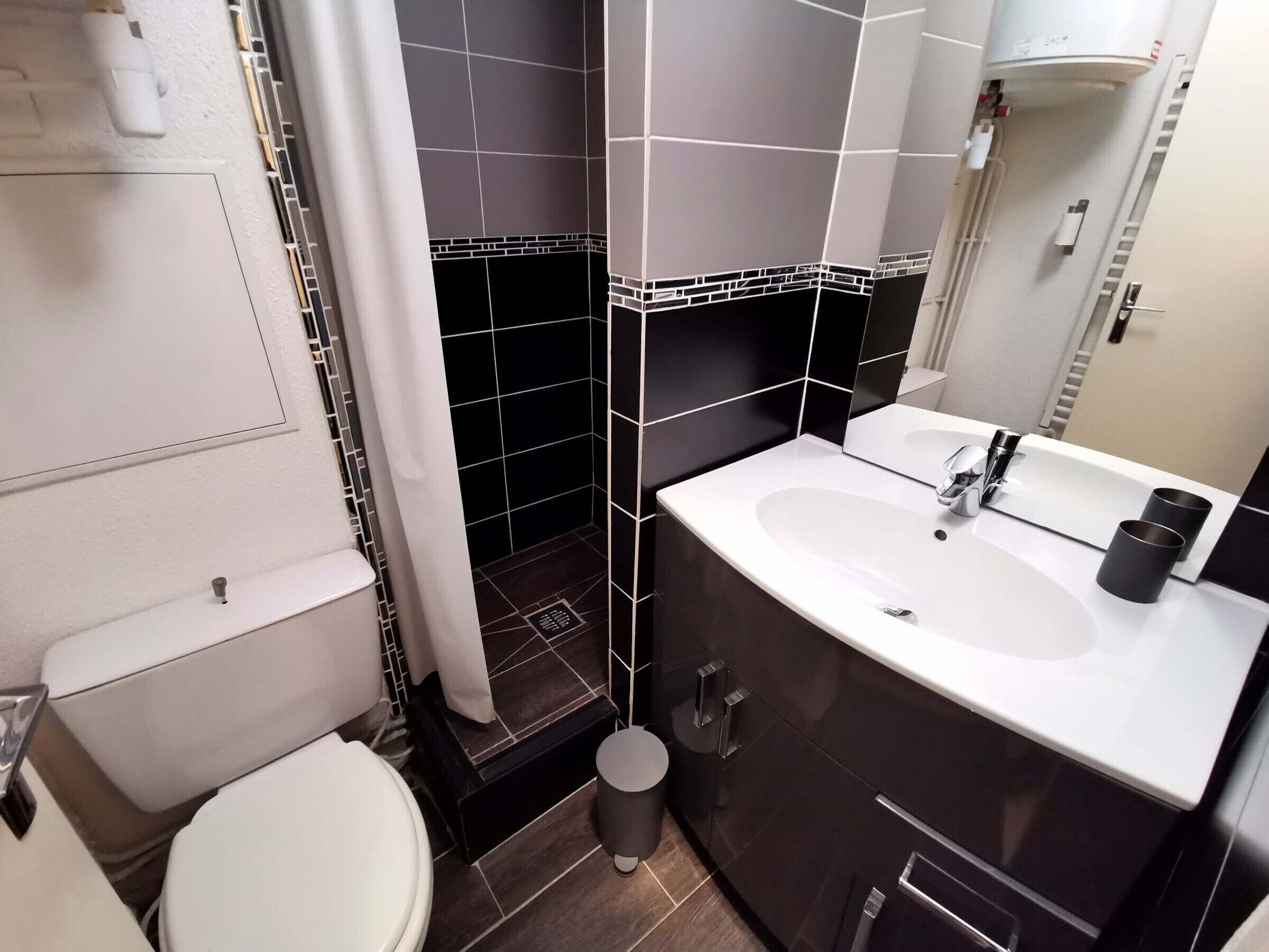 Un baño con inodoro blanco, lavabo con espejo y zona de ducha con azulejos negros.