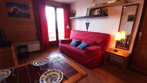 A cozy living room with a red sofa, a dining table set with plates and cutlery, a wooden floor, and a window with red curtains. - Apartment in Adrets-Prapoutel, 2 rooms, balcony, close to slopes and shops (Les Adrets-Prapoutel)