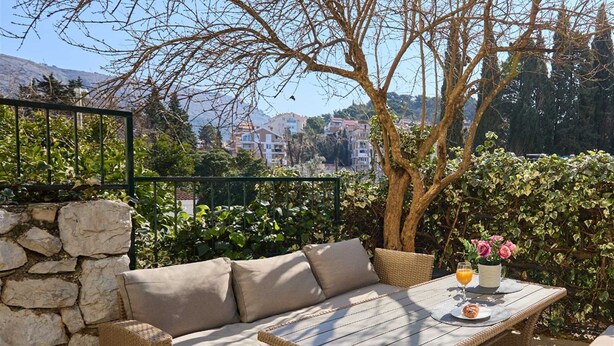 Un espacio al aire libre con sillones y una mesa de mimbre, rodeado de follaje y un muro de piedra.