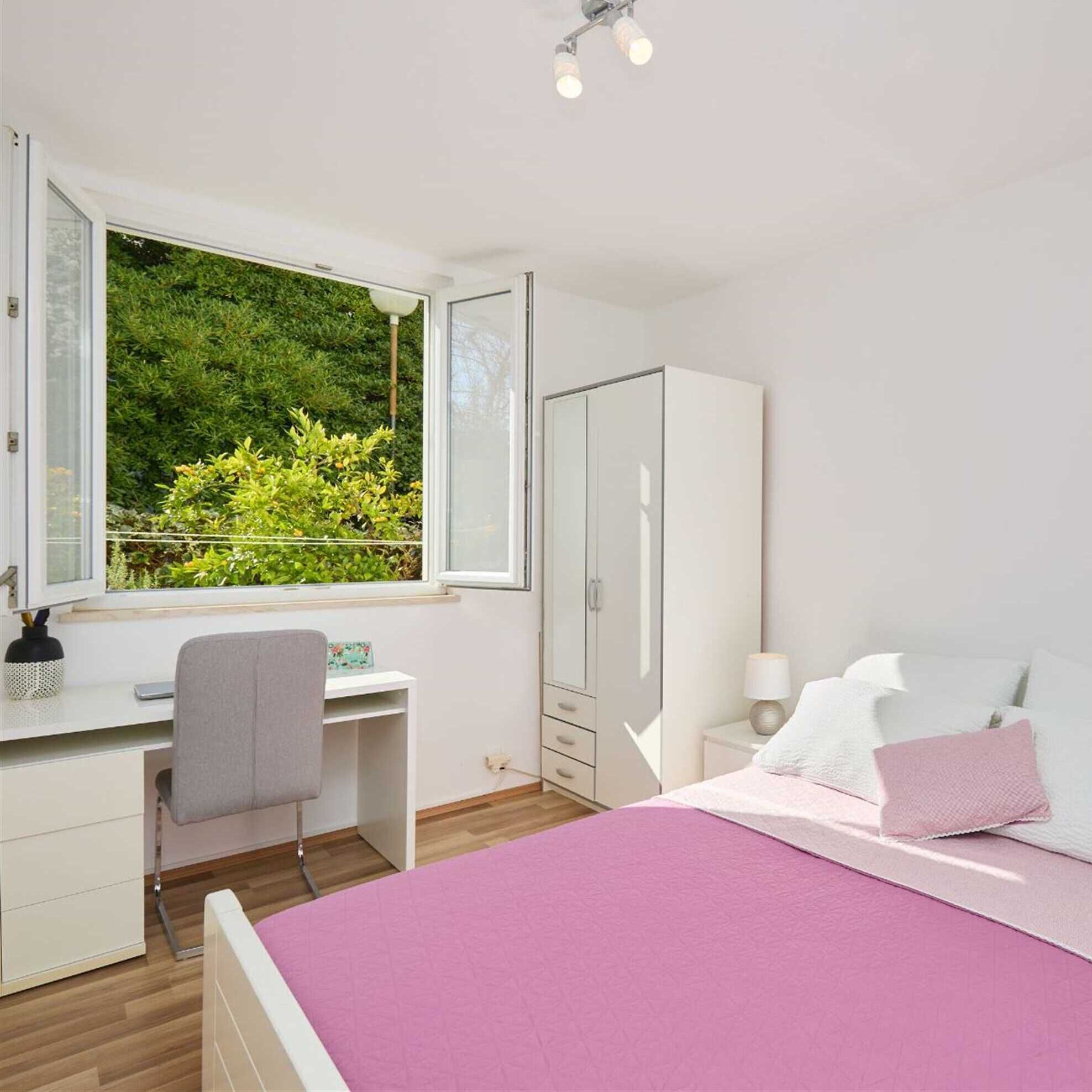 A bedroom with a bed, a desk, a chair, a wardrobe, and a window with a view of greenery.