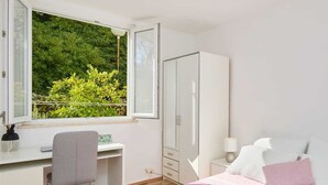 Un dormitorio con cama, escritorio, silla, armario y ventana con vistas a la vegetación.