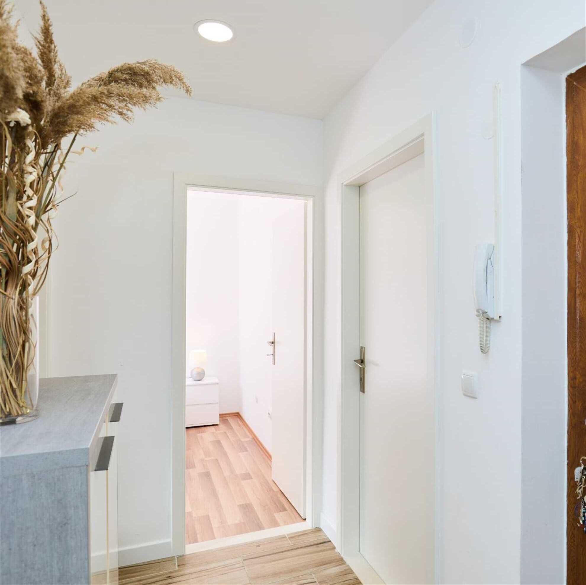 A hallway with a wooden floor, white walls, and a door leading to a room with a bed and a lamp.