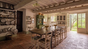 A rustic kitchen with a wooden table and chairs, a hanging pot rack, and a view of the garden through an open door.