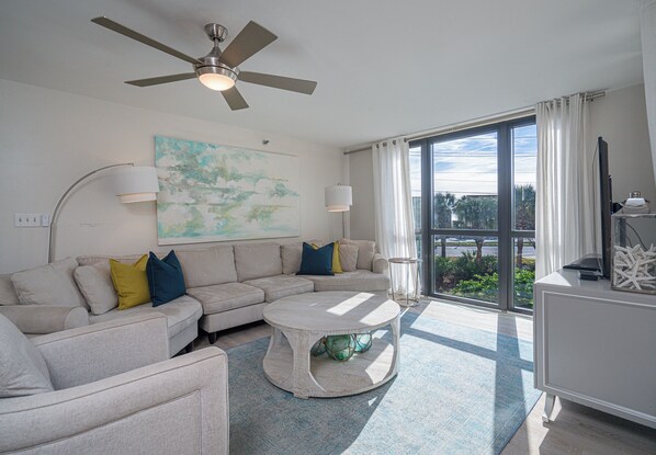 A modern living room with a sectional sofa, a round coffee table, a ceiling fan, and a large window with curtains. - Seafoam Shore- Sleeps 12! (Destin)