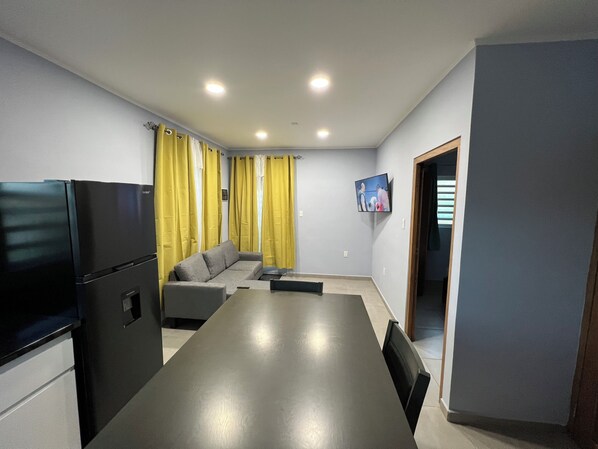 A compact living space with a kitchenette, a sofa, a dining table, and a TV mounted on the wall.
