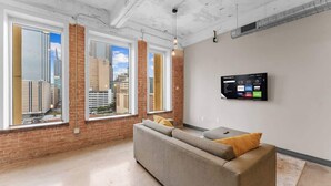 A modern living room with large windows, a grey sofa, and a mounted TV displaying streaming services.