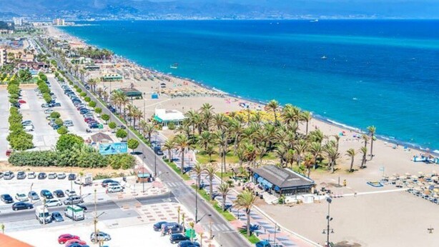 Un paysage urbain côtier avec une plage, un parking et une route bordée de palmiers.