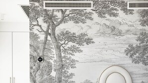 Une chambre avec un grand motif d’arbre peint sur le mur, un lit blanc avec une literie grise, une table de chevet avec une lampe et une armoire blanche.