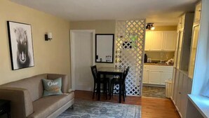 Uma sala de estar com sofá, tapete estampado, área de jantar com mesa e cadeiras, e cozinha com armários e radiador.