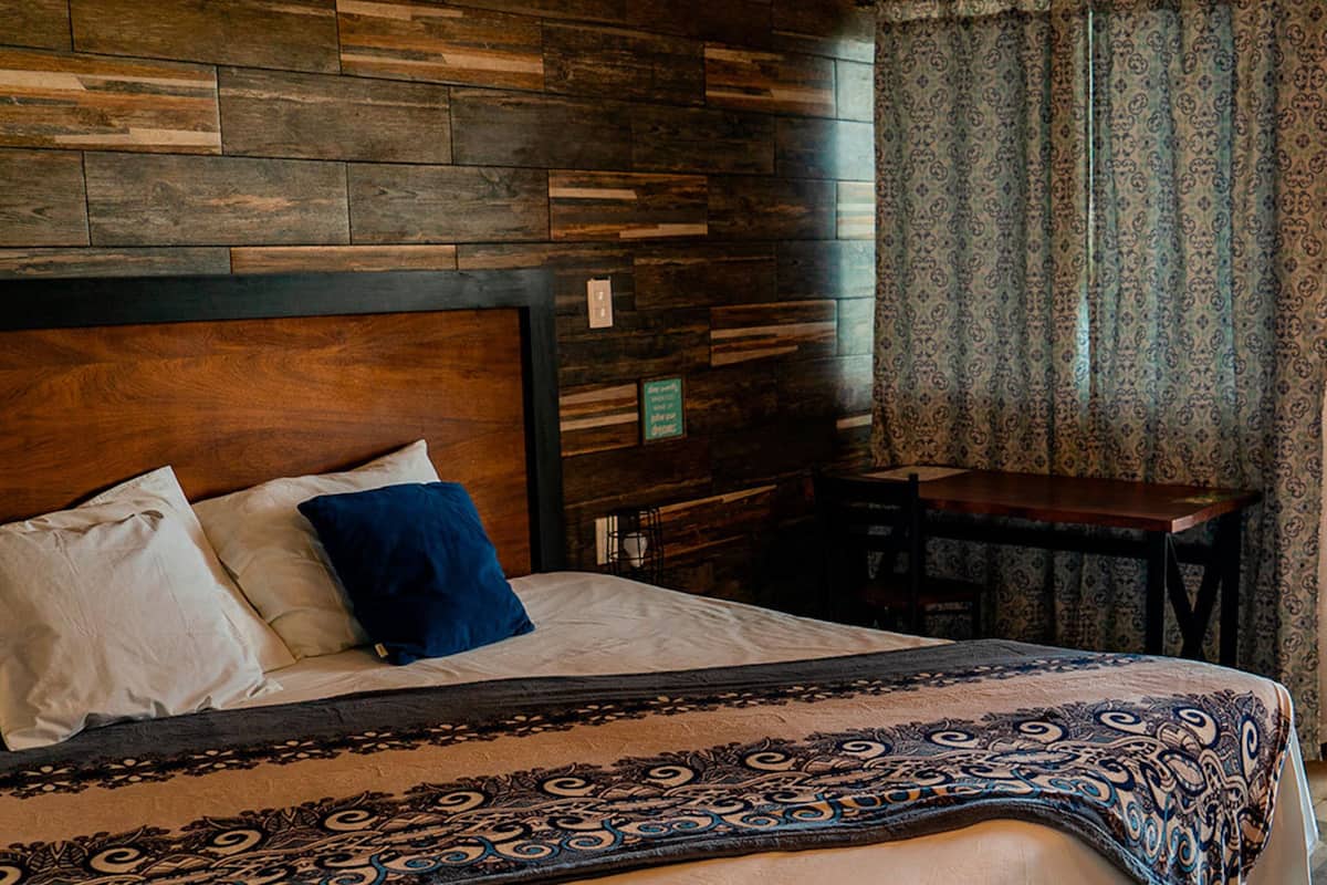 A hotel room with a wooden headboard, a bed with white and blue pillows, a patterned throw, a small table, and patterned curtains.