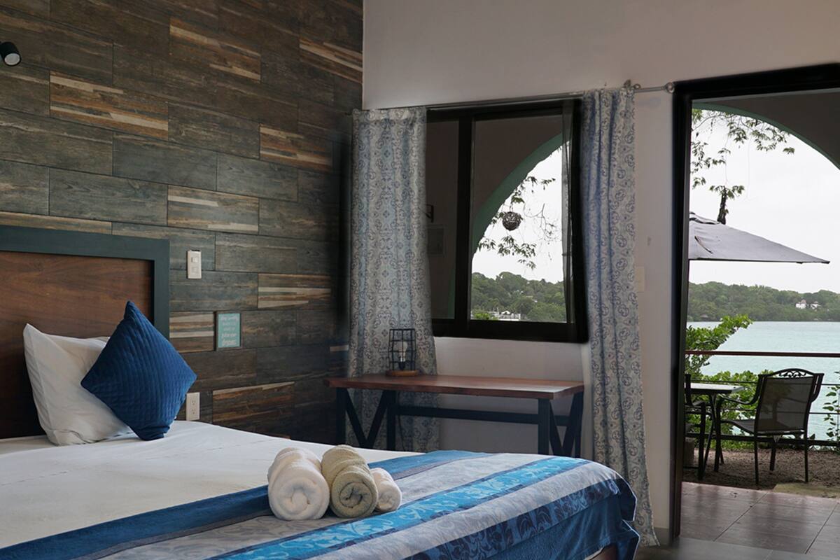 A bedroom with a wooden headboard, a bed with blue bedding, a bedside table, and a view of a balcony with outdoor furniture.