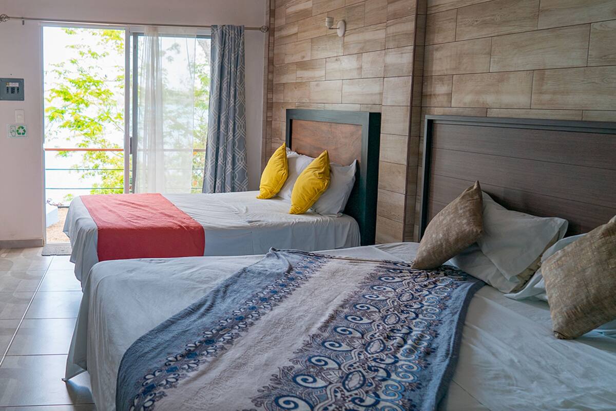 A hotel room with two beds, a wooden wall, a large window with a view, and a ceiling light.