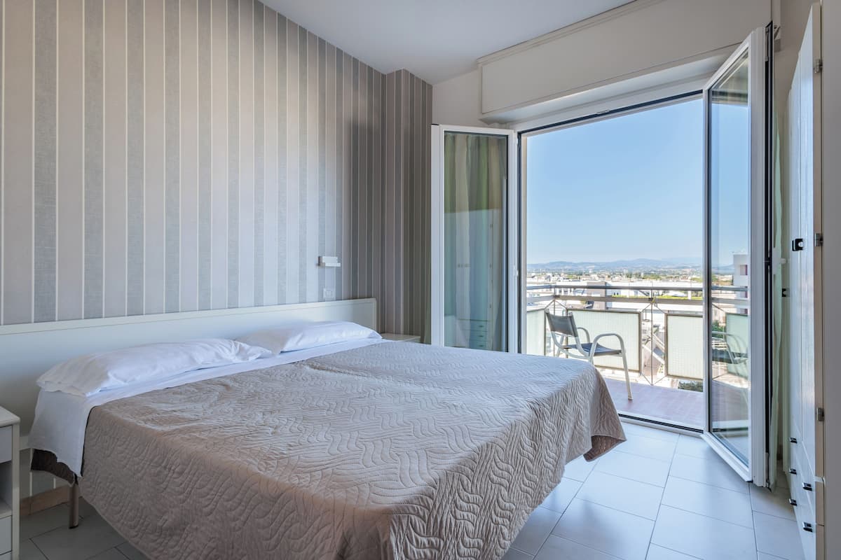 Habitación de hotel con cama, mesita de noche, puerta de cristal corredera a un balcón y vistas a la ciudad.