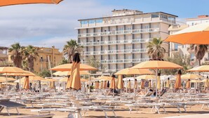 A beach with rows of sun loungers and orange umbrellas, a building in the background, and palm trees. - Hotel Centrale Miramare (Rimini)
