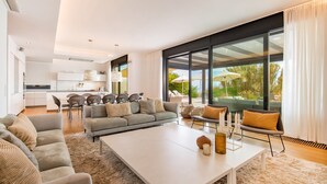 A modern living room with a large white coffee table, grey sofas, and a dining area with chairs. - Villa Dune Costabella (Marbella)