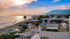 A coastal hotel with a pool, sun loungers, and a view of the beach and mountains.