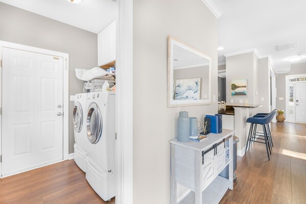 A laundry room with a washing machine, a drying rack, and a small white cabinet with a mirror and a painting.