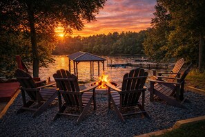 A lakeside seating area with Adirondack chairs around a fire pit, a gazebo, and a dock in the background.