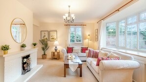 A cozy living room with a fireplace, a sofa set, a coffee table, and large windows offering a view of the garden. - The perfect larger family sized home with vast, lush, enclosed garden. (Trunch)