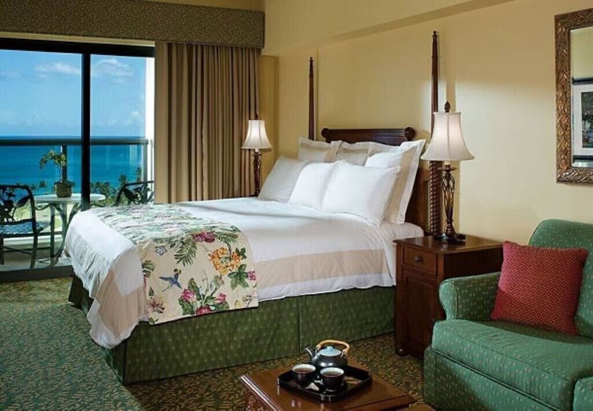 Een hotelkamer met een bed, een groene fauteuil, een nachtkastje en uitzicht op de oceaan.