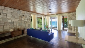 A spacious room with a stone wall, a blue sofa, a small table with a lamp, and a view of the ocean through large windows. - One Bedroom Sea View Villa, Frenchman's Cove Resort (Drapers)
