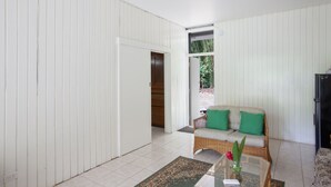 A room with a white paneled wall, a wicker sofa with green cushions, a small table with a vase, and a door leading to another room.