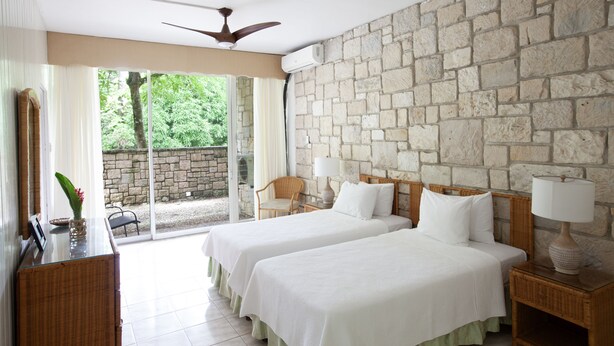 A hotel room with two beds, a stone wall, a ceiling fan, and a view of greenery outside.
