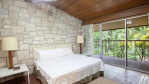 A bedroom with a stone wall, a large bed, two bedside tables with lamps, and a view of greenery through a sliding glass door. - Two Bedroom Garden View Villa, Frenchman's Cove Resort (Drapers)