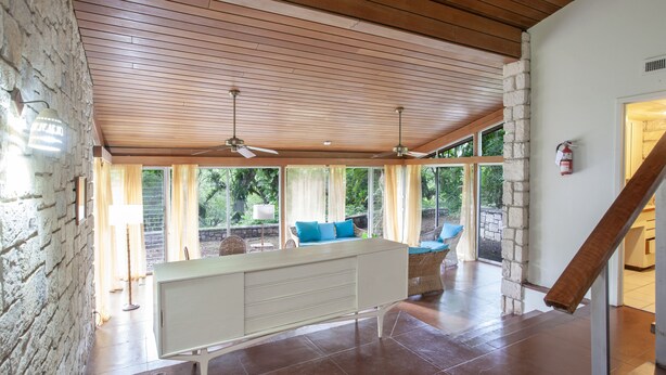A spacious living area with a wooden ceiling, large windows, a white entertainment unit, and a stone wall. - Two Bedroom Garden View Villa, Frenchman's Cove Resort (Drapers)