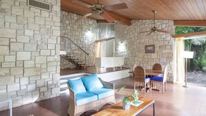 A spacious living area with stone walls, a wooden ceiling, a sofa with blue cushions, a dining table with chairs, and a staircase leading to an upper level. - Two Bedroom Garden View Villa, Frenchman's Cove Resort (Drapers)