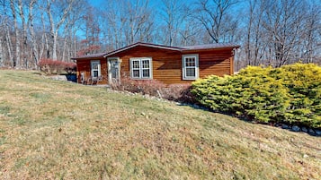 Situated on a grassy slope, this cabin features a central entrance, two ground-floor windows, and a dormer window above.