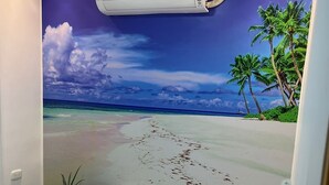 Un dormitorio con un mural de una escena de playa, una cama con sábanas blancas y una mesita de noche pequeña con una planta.