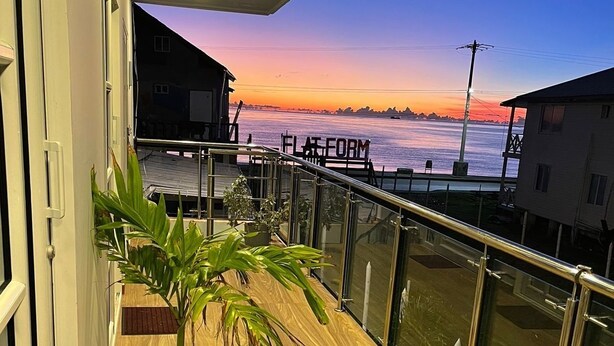 Un balcón con vista al paseo marítimo al atardecer, una planta en maceta y barandilla de cristal.