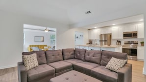 A modern living room with a sectional sofa, a small ottoman, and a kitchen in the background. - Hidden Gem in Pooler (Pooler)