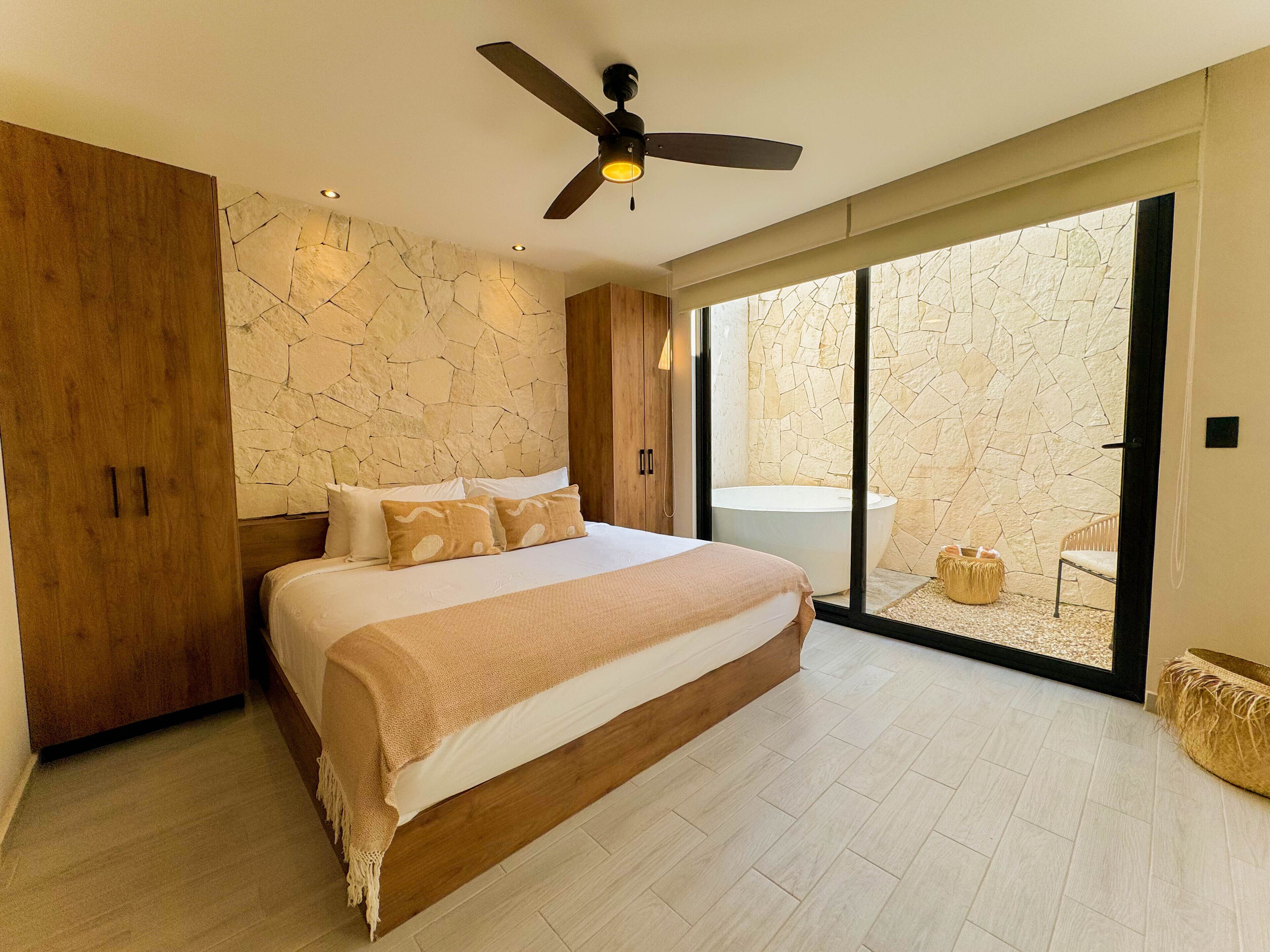 A modern bedroom with a large bed, a ceiling fan, a stone wall, and a view of an outdoor area.
